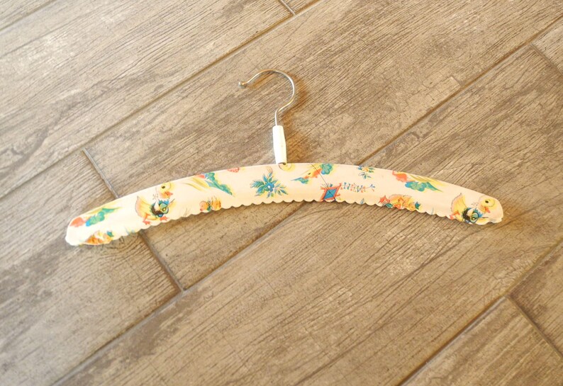 May include: A vintage clothing hanger with a cream-colored base and a scalloped edge. The hanger is decorated with a colorful pattern of birds, flowers, and other whimsical designs. The metal hook is silver.