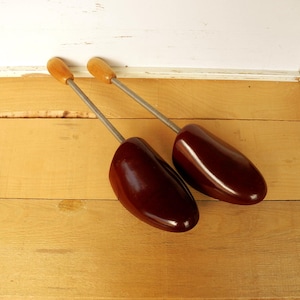 May include: A pair of dark brown shoe trees with wooden handles and metal springs. The shoe trees are made of a glossy material and are designed to maintain the shape of shoes. They are on a light brown wooden surface.