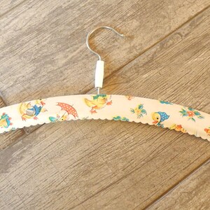 May include: A vintage-style clothes hanger with a white, scalloped edge. The hanger is covered in a fabric featuring a repeating pattern of ducks, umbrellas, and flowers in yellow, blue, and red. The hook is silver.