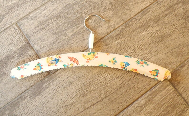 May include: A vintage-style clothes hanger with a white, scalloped edge. The hanger is covered in a fabric featuring a repeating pattern of ducks, umbrellas, and flowers in yellow, blue, and red. The hook is silver.