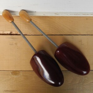 May include: Two vintage shoe trees. The shoe trees have a dark burgundy-coloured shoe form with a wooden handle and a metal spring. The shoe trees are on a light brown wooden surface.
