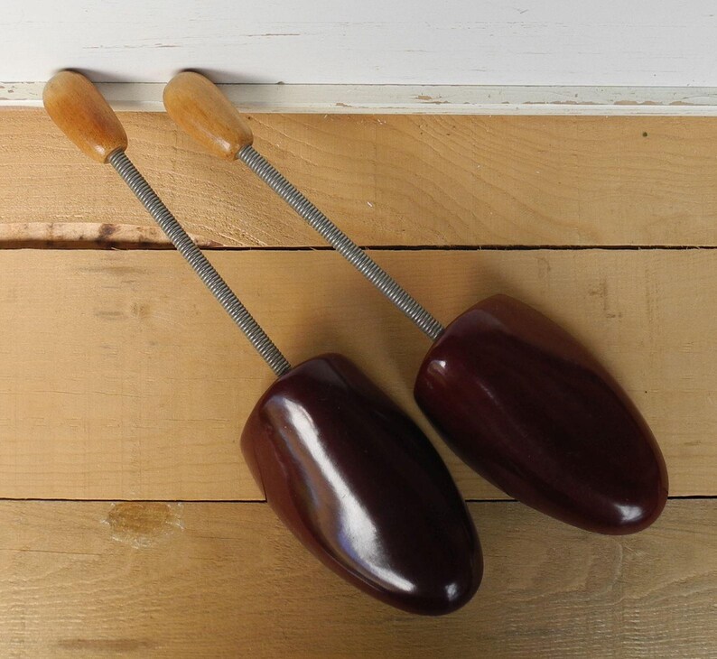 May include: Two vintage shoe trees. The shoe trees have a dark burgundy-coloured shoe form with a wooden handle and a metal spring. The shoe trees are on a light brown wooden surface.