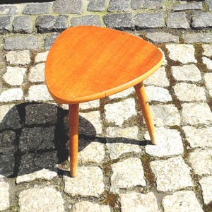 Könnte beinhalten: Ein kleiner, dreibeiniger Holzschemel mit dreieckiger Sitzfläche. Der Hocker ist aus hellem Holz mit einer warmen, orangebraunen Oberfläche gefertigt. Die Beine sind gleichmäßig verteilt und nach außen abgewinkelt. Der Hocker ist etwa 30 cm hoch.
