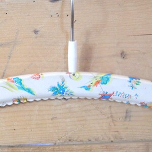 May include: Vintage white metal clothes hanger with a decorative scalloped edge. The hanger is adorned with a colorful pattern of birds and floral designs. It has a white plastic hook and a metal rod.