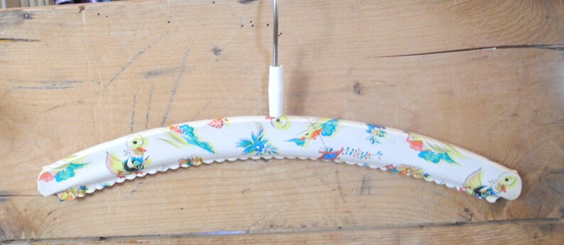 May include: Vintage white metal clothes hanger with a decorative scalloped edge. The hanger is adorned with a colorful pattern of birds and floral designs. It has a white plastic hook and a metal rod.