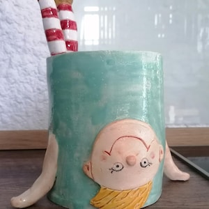 Op de afbeelding: Een grillige keramische plantenbak, cilindrisch van vorm en turquoise geschilderd. Het heeft een gezicht en armen, met benen in rood-wit gestreepte kousen. Dit unieke ontwerp is geschikt voor kleine planten.