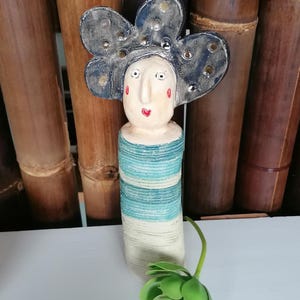 Könnte beinhalten: Skurrile Keramikfigur mit blütenförmigem Kopfschmuck und gestreiftem Körper in Blau- und Weißtönen. Das Gesicht hat rote Akzente. Eine grüne Blume befindet sich im Vordergrund. Die Figur ist ein dekoratives Objekt.