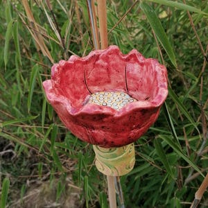 Op de afbeelding: Een handgemaakte, rode keramische vogelvoeder in de vorm van een bloem. De bloem heeft een getextureerd oppervlak en een geel en wit interieur. Het is gemonteerd op een houten stok met een groen en geel patroon, tegen een achtergrond van bamboe.