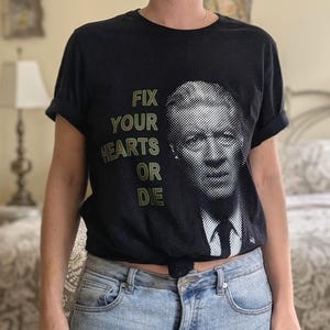 May include: A black short-sleeved t-shirt featuring a halftone portrait of a man in a suit and tie. To the left of the portrait, the text "FIX YOUR HEARTS OR DIE" is printed in a stacked, olive green, block-style font on the front of the shirt.