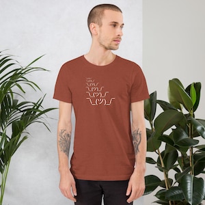May include: A rust-colored short-sleeve t-shirt with a white graphic of a shrug emoji. The graphic is stacked in a descending order. The shirt is made of a soft material.