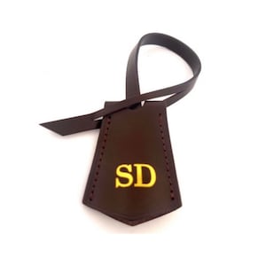 May include: A dark brown leather luggage tag with the initials "SD" in gold lettering. The tag has a loop for attaching to a suitcase or bag and is stitched with red thread.