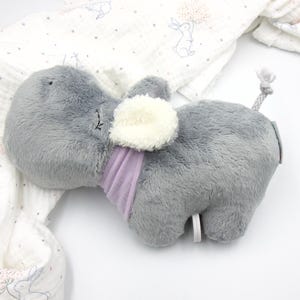 Op de afbeelding: Een grijs pluche olifantenspeelgoed met een paarse sjaal en een witte, pluche staart. De olifant ligt op zijn zij met zijn hoofd rustend op een wit deken.