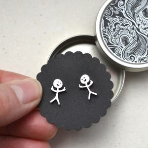 Könnte beinhalten: Ein Paar silberne Ohrringe in Form von Strichmännchen auf einer schwarzen, geschwungenen Karte. Die Ohrringe stellen eine Person mit erhobenen Armen dar. Die Karte befindet sich in einer offenen silbernen Dose mit einem dekorativen schwarz-weißen Blumendesign.