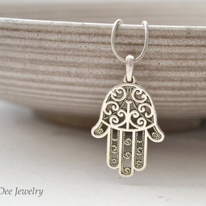 Può includere: Una collana con pendente a mano di Hamsa color argento. Il pendente presenta un design traforato con motivi intricati ed è sospeso a una delicata catena. La collana è mostrata su uno sfondo neutro.