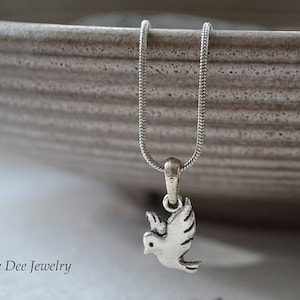 Op de afbeelding: Een zilverkleurige ketting met een duifhanger. De duif wordt afgebeeld met uitgespreide vleugels en de ketting heeft een fijne ketting. De achtergrond is wazig, neutraal, waardoor de sieraden worden benadrukt. De merknaam "Jane Dee Jewelry" is zichtbaar.