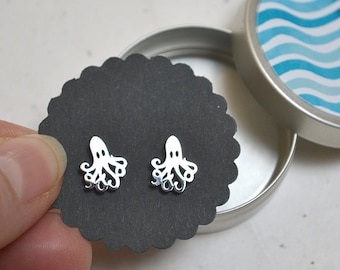 May include: A pair of silver octopus stud earrings displayed on a black scalloped card. The earrings are in a small, round, silver tin with a blue and white wave pattern on the lid. The earrings are small and delicate.