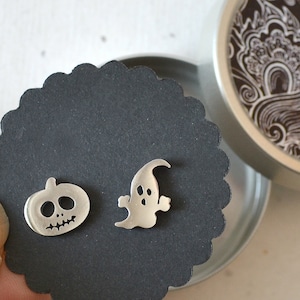 Könnte beinhalten: Silberne Halloween-Ohrstecker. Ein Ohrring ist ein Kürbis mit geschnitztem Gesicht, der andere ein Geist. Die Ohrringe werden auf einer schwarzen, wellenförmigen Karte in einer dekorativen Dose präsentiert.