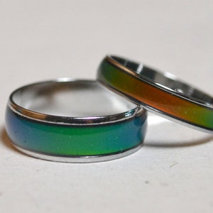 Op de afbeelding: Twee zilveren stemmingringen met een kleurrijke, iriserende band. De bovenste ring heeft een groene, oranje en gele band. De onderste ring heeft een blauwe, groene en gele band.