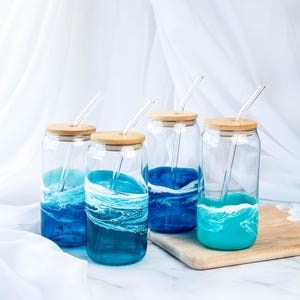 K&ouml;nnte beinhalten: Vier gl&auml;serne Trinkgl&auml;ser mit Holzdeckeln und Glasstrohhalmen. Jedes Glas hat ein anderes blaues und wei&szlig;es Meereswellendesign.