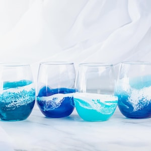 Puede incluir: Cuatro copas de vino de vidrio transparente con diseños de olas de mar azules y blancas. Las copas están dispuestas en fila sobre una superficie blanca.