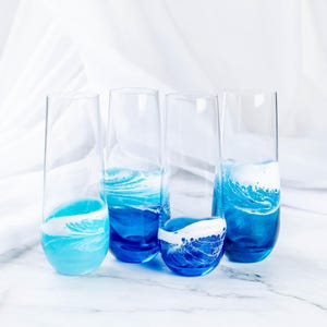 Può includere: Quattro flûte da champagne in vetro trasparente con disegni astratti blu e bianchi che ricordano le onde dell'oceano. I bicchieri sono disposti in fila su una superficie di marmo bianco.