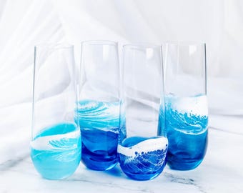 Könnte beinhalten: Vier klare Champagnerflöten aus Glas mit blauen und weißen abstrakten Mustern, die an Meereswellen erinnern. Die Gläser sind in einer Reihe auf einer weißen Marmorfläche angeordnet.