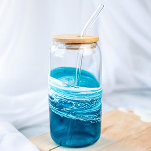 K&ouml;nnte beinhalten: Eine Glaswasserflasche mit einem blauen und wei&szlig;en Wirbelmuster und einem Holzdeckel. Ein Glasstrohhalm befindet sich in der Flasche.