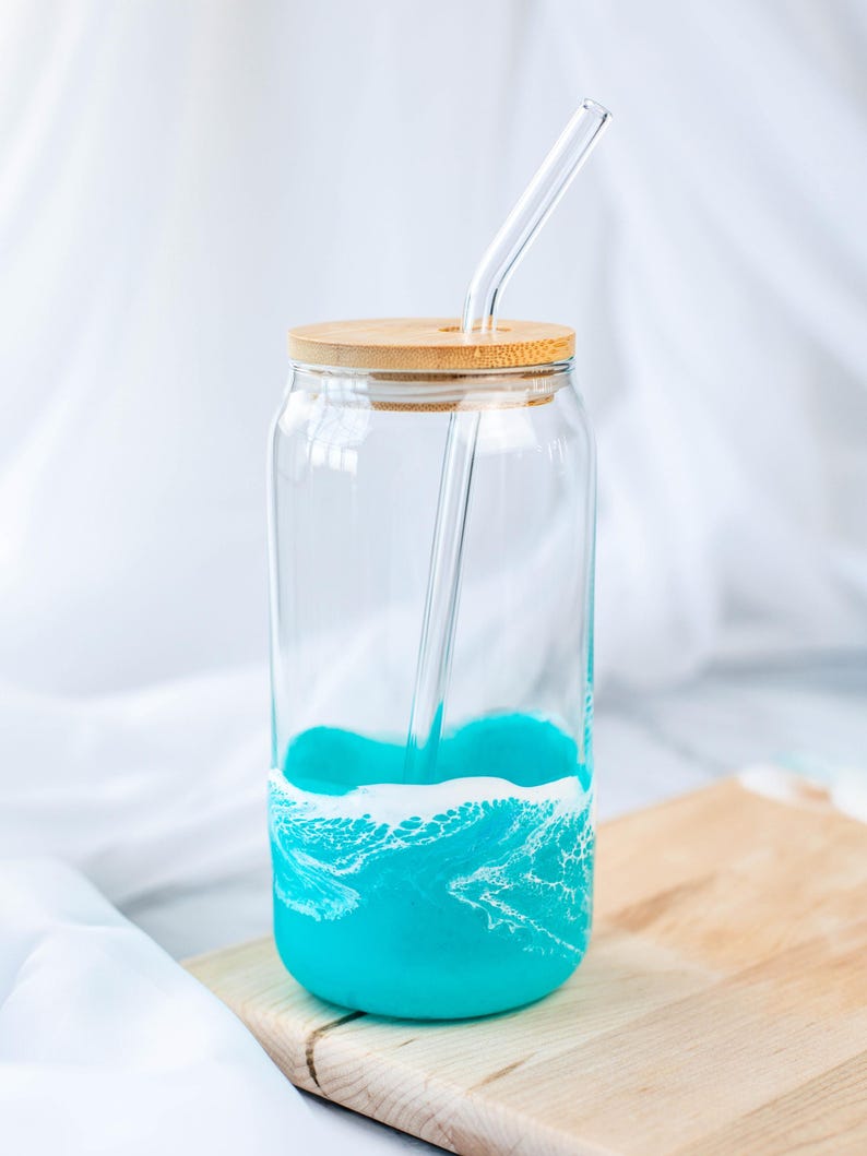 K&ouml;nnte beinhalten: Ein Glas-Trinkglas mit einem t&uuml;rkisblauen und wei&szlig;en Wellendesign. Das Glas hat einen Holzdeckel und einen Glasstrohhalm.