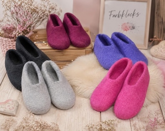 May include: Four pairs of felted wool slippers in dark gray, light gray, magenta, and purple. The slippers have a rounded toe, a low-cut opening, and a textured wool finish. A sign with the text "Farbflecks" is visible in the background.