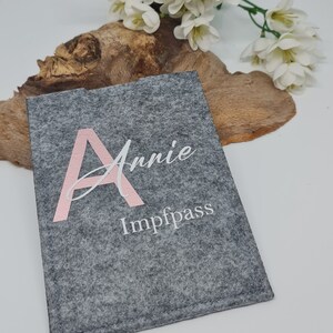 Könnte beinhalten: Ein grauer Filzbeutel mit einem rosafarbenen Buchstaben A und dem Namen "Annie" sowie dem Wort "Impfpass" in weißer Schrift.