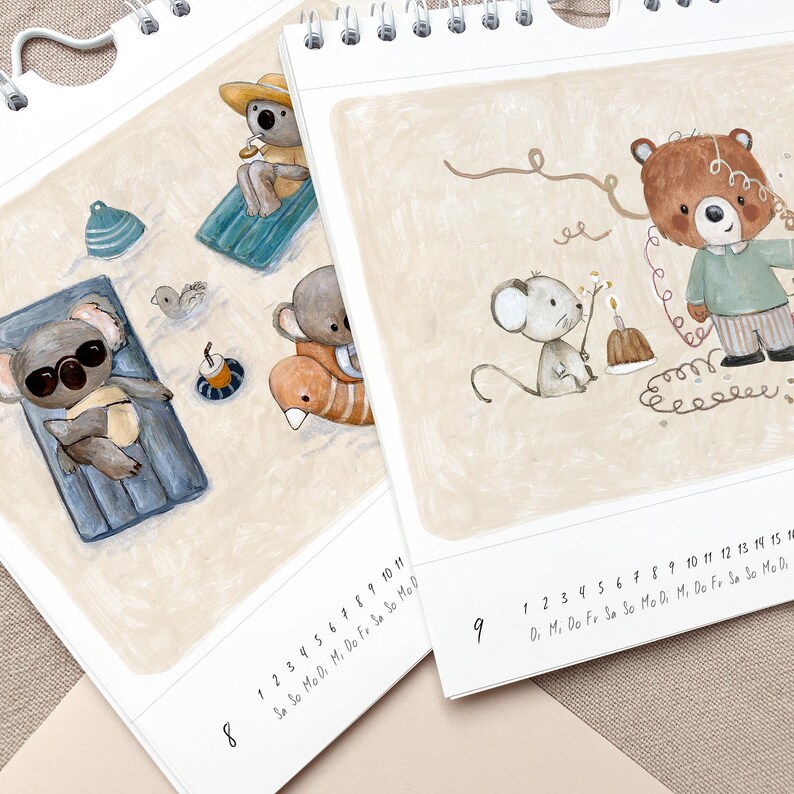 May include: Two open spiral-bound calendars with illustrations of animals. One calendar shows koalas on a beach, the other shows a bear and mouse. The calendars have dates at the bottom. The background is beige.