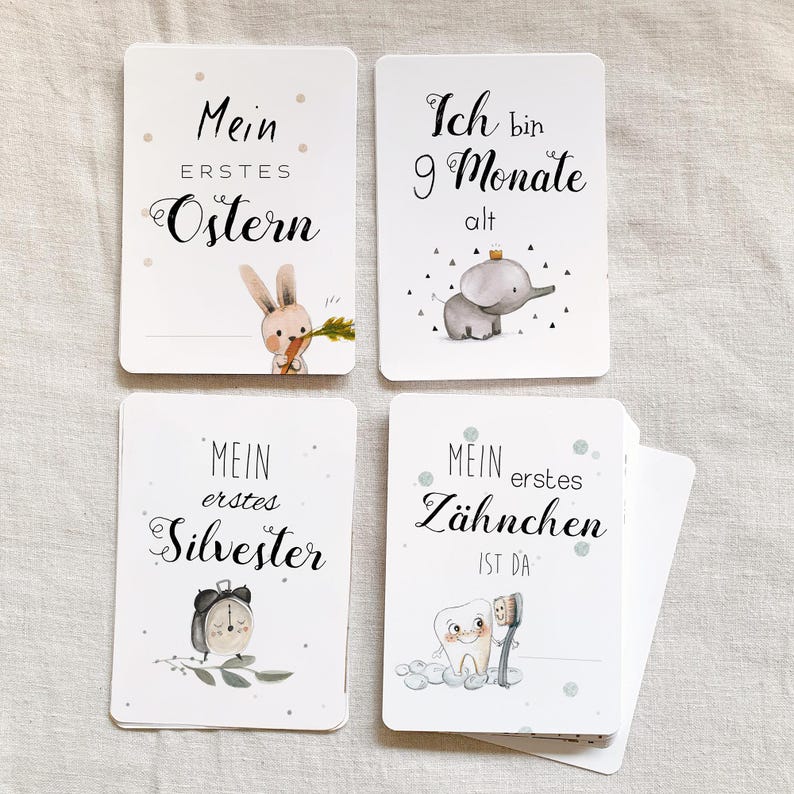 May include: Four white cards with black text and illustrations. The cards feature milestones for a baby, such as "My First Easter", "My First Tooth", "My First New Year's Eve", and "I am 9 months old".