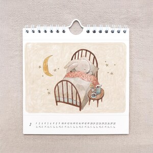 May include: A spiral-bound calendar featuring an illustration of a sleeping bunny in a bed. The bunny is covered with a pink blanket, and a small table with a cup and saucer sits beside the bed. A crescent moon and stars are in the background.