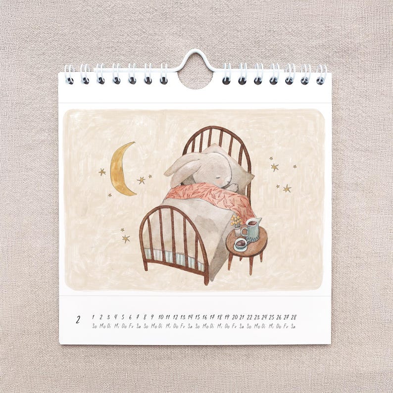 May include: A spiral-bound calendar featuring an illustration of a sleeping bunny in a bed. The bunny is covered with a pink blanket, and a small table with a cup and saucer sits beside the bed. A crescent moon and stars are in the background.