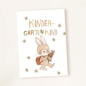 Könnte beinhalten: Aquarell-Illustration eines lächelnden Hasen mit Rucksack, der einen kleineren Hasen hält. Der Text "Kindergarten Kind" steht in bunten Buchstaben über dem Hasen, mit Sternen-Akzenten. Das Kunstwerk befindet sich auf weißem Hintergrund.