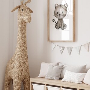 May include: A light brown woven giraffe toy stands next to a white wooden storage bench with four drawers. The bench has three gray and white pillows on top. A framed print of a gray and white cartoon cat is hanging on the wall above the bench. A white and gray bunting is hanging above the print.