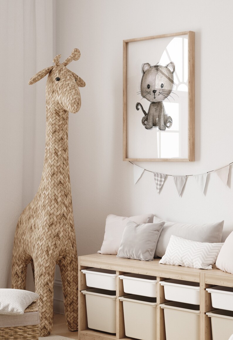 May include: A light brown woven giraffe toy stands next to a white wooden storage bench with four drawers. The bench has three gray and white pillows on top. A framed print of a gray and white cartoon cat is hanging on the wall above the bench. A white and gray bunting is hanging above the print.