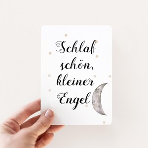 May include: White card with black text that reads "Schlaf sch&ouml;n, kleiner Engel" and a crescent moon illustration. The card has small gold glitter dots scattered across the surface.