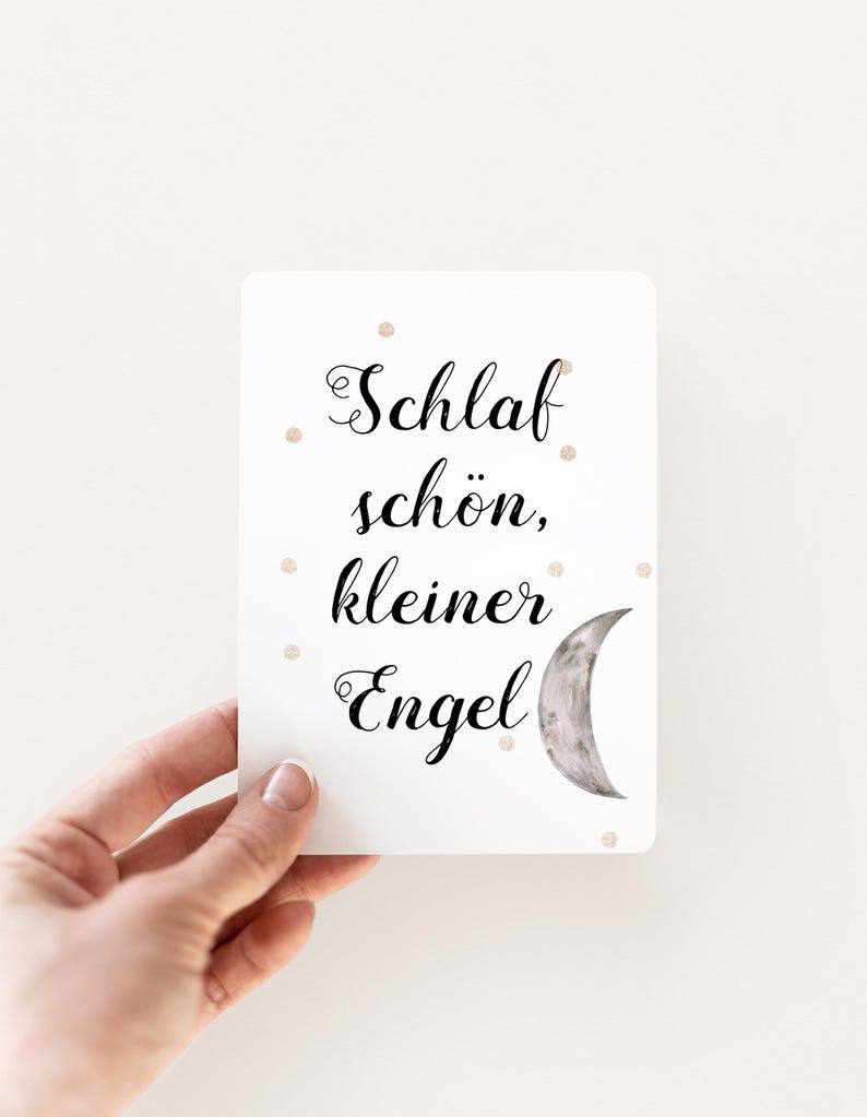 May include: White card with black text that reads "Schlaf sch&ouml;n, kleiner Engel" and a crescent moon illustration. The card has small gold glitter dots scattered across the surface.