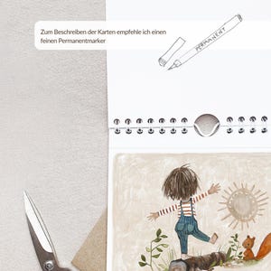 May include: A spiral-bound calendar with a watercolor illustration of a child balancing on a log. The calendar has a white background with the word "PERMANENT" and German text. A pair of scissors is visible.