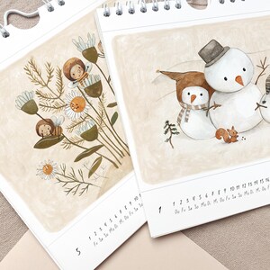 May include: Two spiral-bound calendars with illustrations. One features a bouquet of flowers with bee-like characters. The other shows a snowman and a smaller snowman with a squirrel. The calendars have a neutral color palette with the days of the week printed at the bottom.