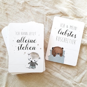 May include: Two white cards with black text and illustrations. The first card says "Ich kann jetzt alleine stehen" and features a small otter wearing a blue shirt. The second card says "Ich & mein liebstes Kuscheltier" and features a brown bear sleeping in a blue blanket.