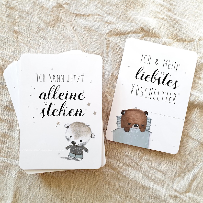 May include: Two white cards with black text and illustrations. The first card says "Ich kann jetzt alleine stehen" and features a small otter wearing a blue shirt. The second card says "Ich & mein liebstes Kuscheltier" and features a brown bear sleeping in a blue blanket.