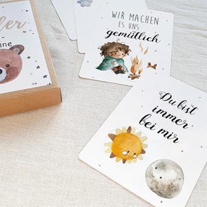 May include: A set of cards with illustrations and text in German. The cards feature a girl sitting by a campfire, a sun and moon, and a bear. The text on the cards reads "Wir machen es uns gem&uuml;tlich" and "Du bist immer bei mir".