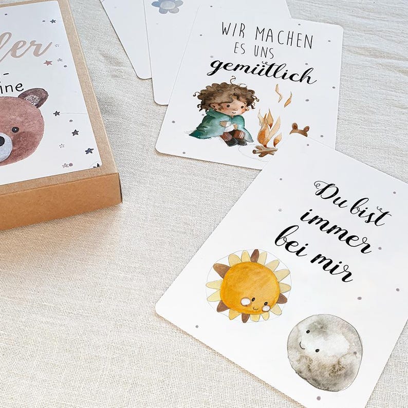May include: A set of cards with illustrations and text in German. The cards feature a girl sitting by a campfire, a sun and moon, and a bear. The text on the cards reads "Wir machen es uns gem&uuml;tlich" and "Du bist immer bei mir".