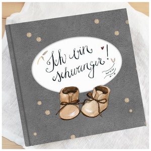May include: A gray baby book cover with a white oval containing the text "Ich bin schwanger!" and a small heart. The oval is surrounded by small gold dots. Two brown baby shoes are at the bottom of the cover.