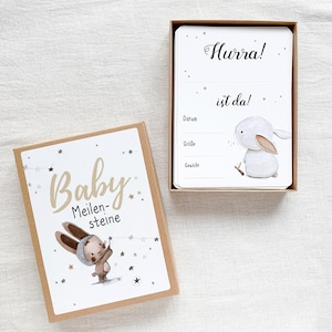 May include: A brown box with a white bunny illustration and the text "Baby Meilensteine" on the front. The box contains a white card with the text "Hurra! ist da!" and spaces to record the date, size, and weight.