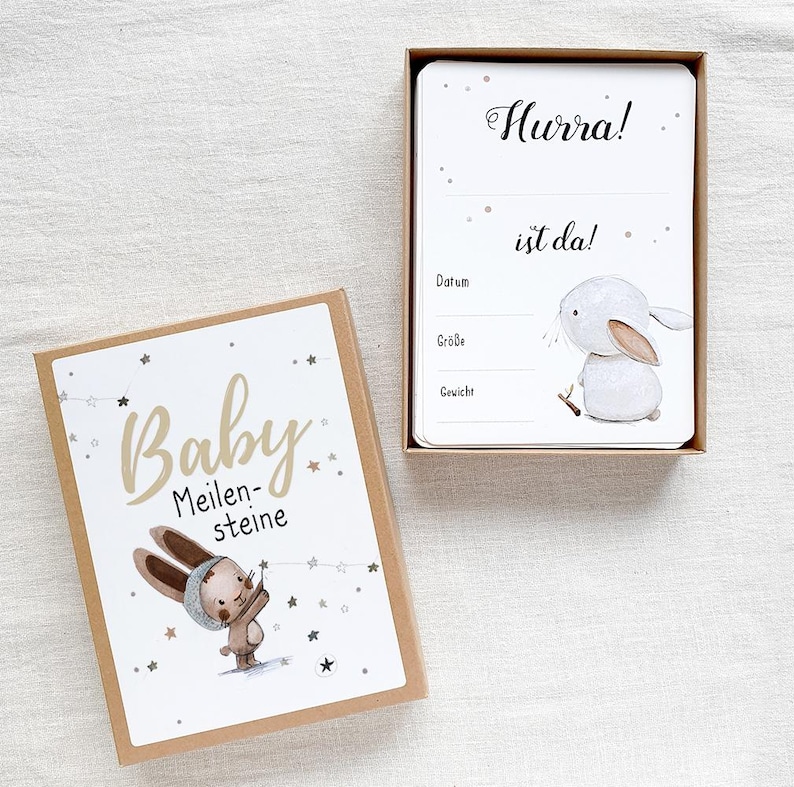 May include: A brown box with a white bunny illustration and the text "Baby Meilensteine" on the front. The box contains a white card with the text "Hurra! ist da!" and spaces to record the date, size, and weight.