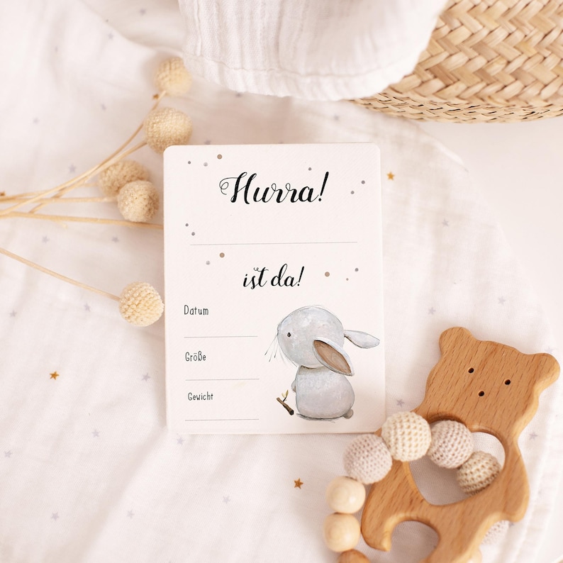 May include: A white card with the text "Hurra! ist da!" and a gray bunny illustration. The card has lines for "Datum", "Gr&ouml;&szlig;e", and "Gewicht".