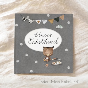 Könnte beinhalten: Eine graue Karte mit einem weißen Oval, das den Text "Unser Enkelkind" in schwarzer Kursivschrift enthält. Ein brauner Bär mit weißem Bauch und einem Pinsel sitzt auf der Karte mit einer Palette an Farben. Die Karte ist mit einer Girlande aus Flaggen und silbernem Glitzer verziert.